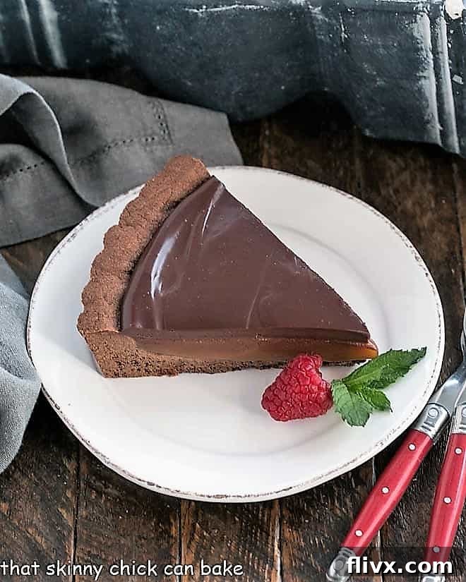 A perfectly sliced piece of Chocolate Caramel Tart, showcasing its distinct layers of chocolate crust, golden caramel, and glossy ganache, served on a pristine white plate.