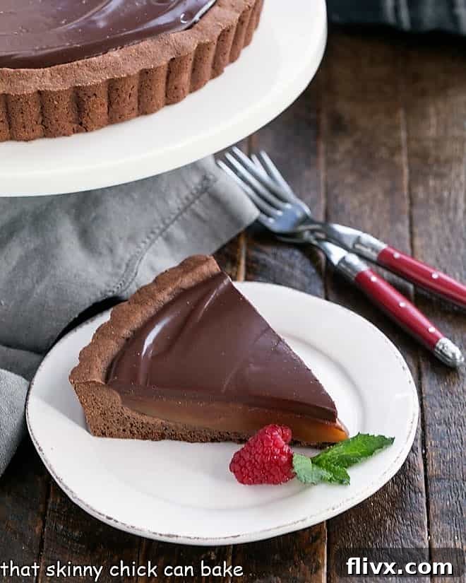 A pristine slice of Chocolate Caramel Tart resting on a white plate, flanked by a dessert stand and two forks, inviting indulgence.
