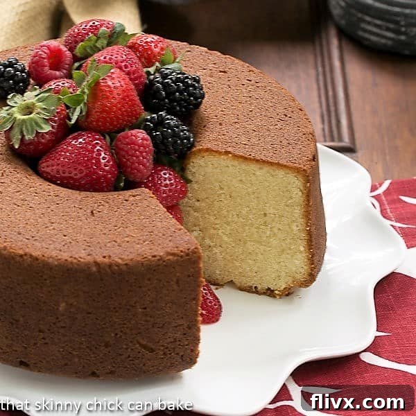 Vanilla Pound Cake - Perfect dessert topped with summer berries or any seasonal topping Whole Vanilla Pound Cake on a white platter with one slice removed, revealing its inviting interior and a pile of fresh berries in the center.