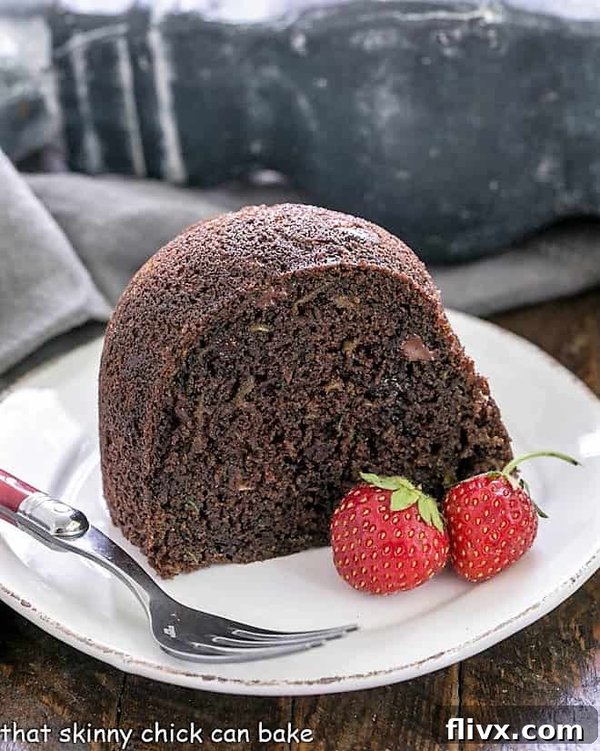 A perfectly portioned slice of Chocolate Zucchini Bundt Cake on a white plate, accompanied by a fork with a red handle and two fresh strawberries, highlighting its moist texture.
