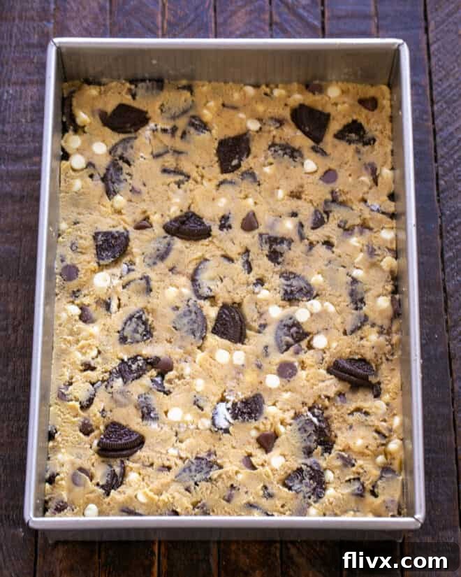 Cookie dough being spread evenly into a baking pan coated with nonstick spray.