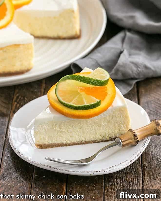 Margarita Cheesecake - An exquisite citrus cheesecake with the flavors of a margarita! A perfectly sliced Margarita Cheesecake on a white dessert plate, elegantly garnished with fresh orange and lime slices. The vibrant green and yellow hues hint at the refreshing citrus flavor within.