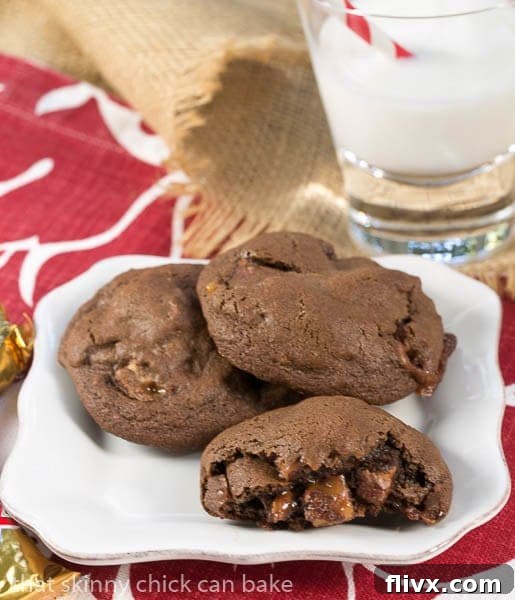 A close-up of a Double Chocolate Twix Cookie on a white plate, with one broken open to reveal the gooey candy-filled interior of caramel and chocolate