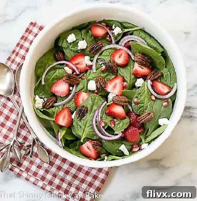 A vibrant Spinach, Strawberry, Pomegranate, Feta Salad served in an elegant white bowl, placed on a classic red and white checked napkin.