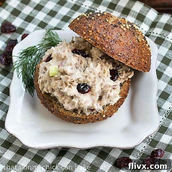 A beautifully presented Tuna, Cranberry, Pecan Salad Sandwich, cut in half, on a pristine white plate, garnished with fresh greens.