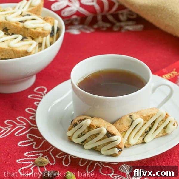 Two cherry pistachio biscotti on a white saucer with a cup of tea, showcasing their festive colors and crunchy texture.
