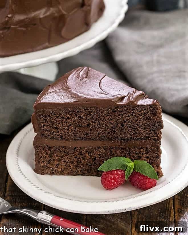 A pristine slice of rich Devil's Food Cake, featuring its dark layers and creamy chocolate frosting, presented on a white dessert plate with a red-handled fork. A vintage cake stand is softly blurred in the background, adding to the elegant setting.