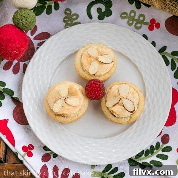 Three beautifully presented Almond Raspberry Tartlets on a white plate, garnished with fresh raspberries.
