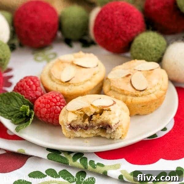 Almond Raspberry Tartlets on a white plate, with one tart cut open to expose its delicious layers of raspberry jam and almond paste filling.
