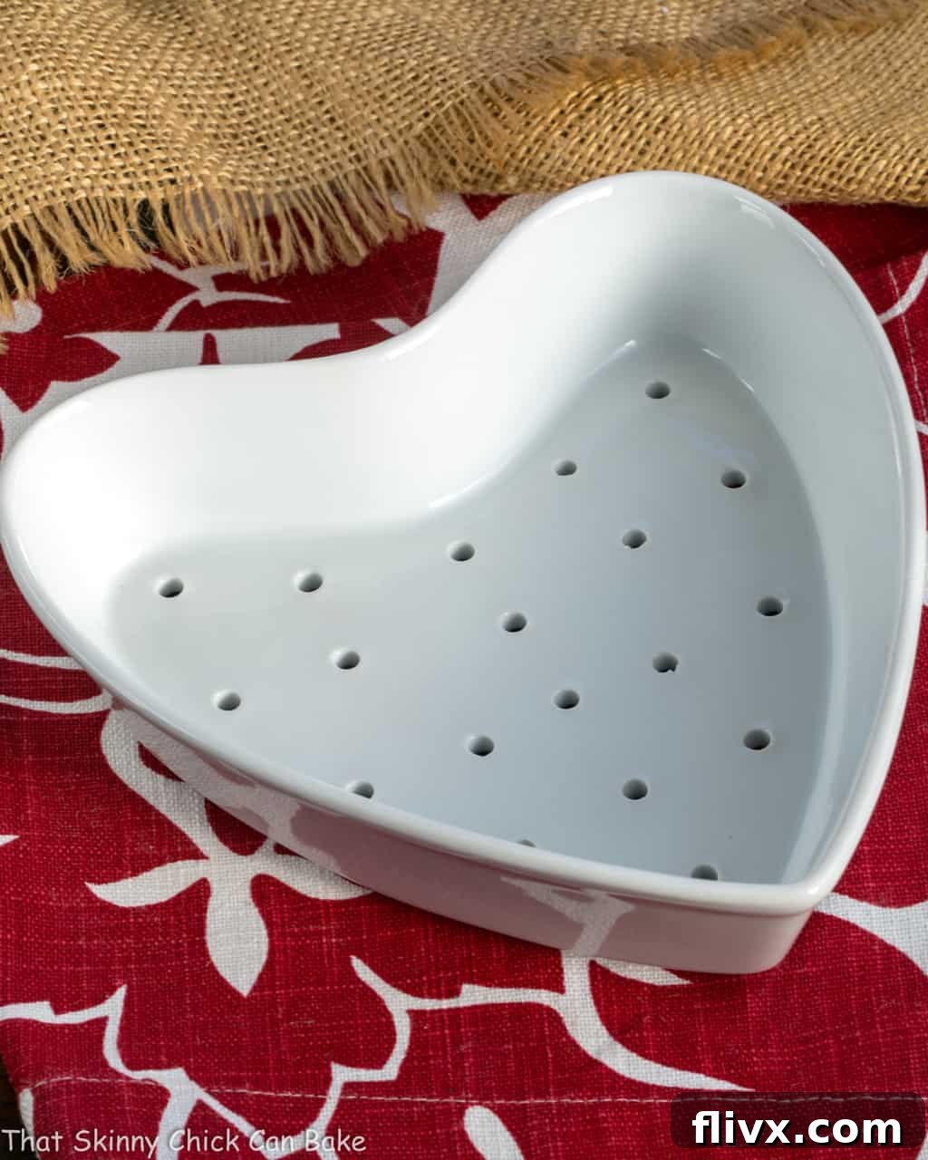 A ceramic heart-shaped Coeur a la Creme mold lined with cheesecloth, resting on a festive red and white striped napkin, ready for the creamy chocolate mixture.