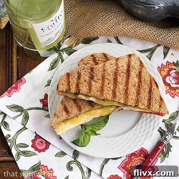 Overhead view of a Chicken Pesto Grilled Cheese sandwich on a white plate with a basil garnish and a red-handled fork, highlighting its golden crust and melted cheese.