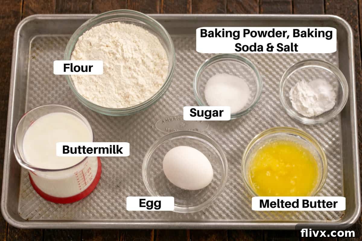 A beautifully arranged metal sheet pan showcasing all the essential ingredients for buttermilk pancakes, each clearly labeled for easy identification: flour, sugar, salt, baking soda, baking powder, egg, buttermilk, and melted butter.