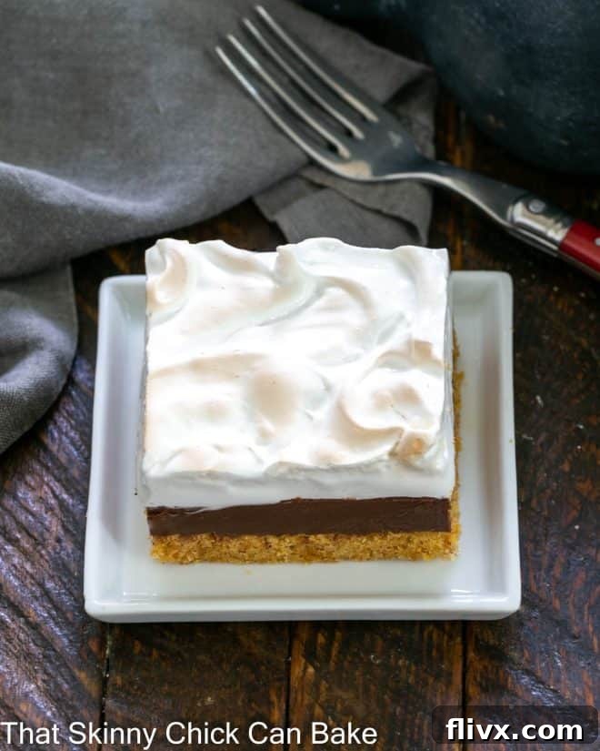 Dreamy Smores Fudge Bars 2 A perfectly portioned S'mores Fudge Bar on a small white square ceramic plate, showcasing its three distinct layers: golden graham cracker crust, dark fudge, and toasted marshmallow meringue. A fork is nearby, ready for a bite.