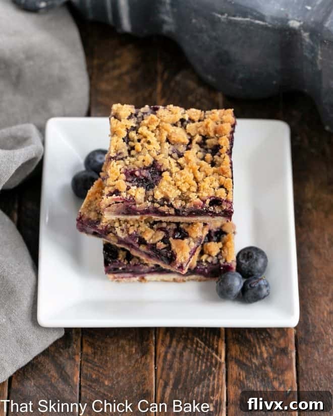 A beautifully stacked trio of golden blueberry streusel bars, garnished with fresh blueberries, on a pristine white plate, ready to be enjoyed.