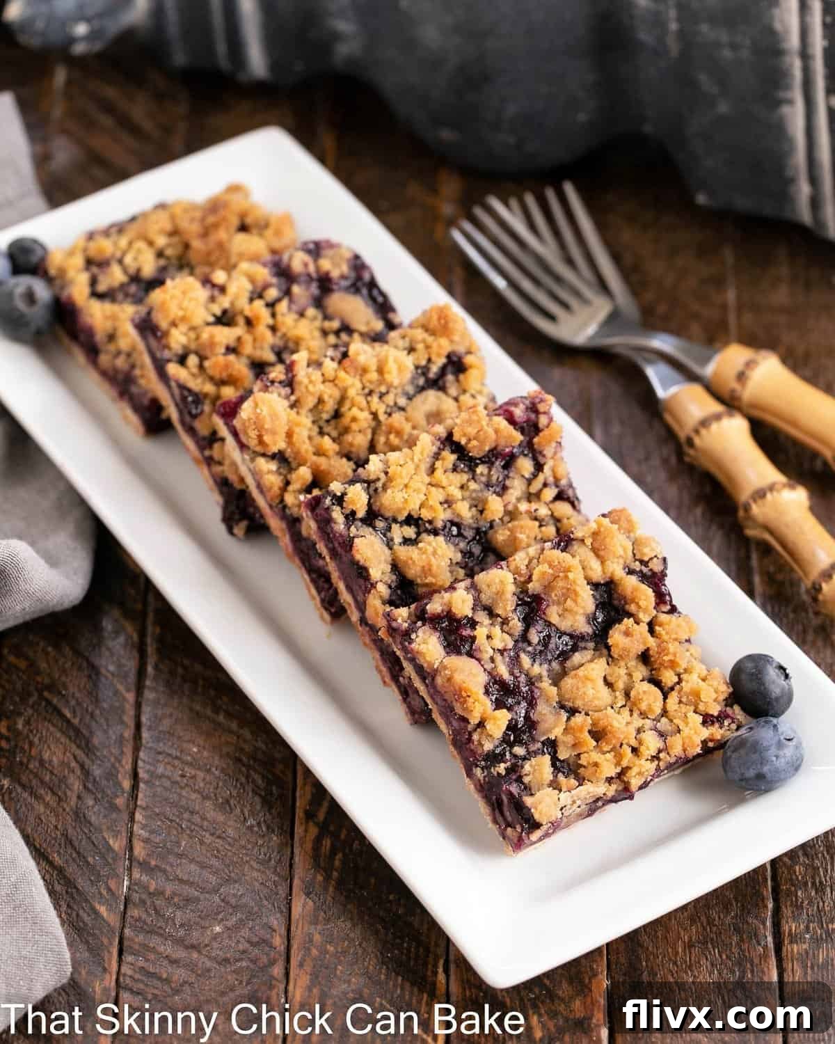 A collection of freshly baked Blueberry Streusel Bars elegantly arranged on a rustic white serving tray, accompanied by two delicate bamboo forks.