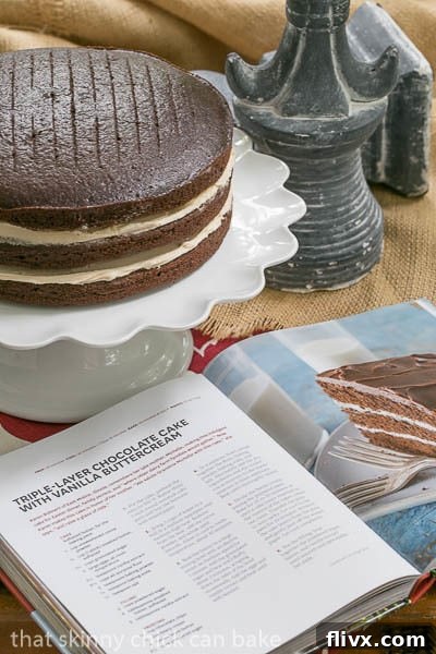 Unfrosted chocolate cake layers with vanilla buttercream filling, stacked on a cake stand, next to an open cookbook, ready for the final touch.
