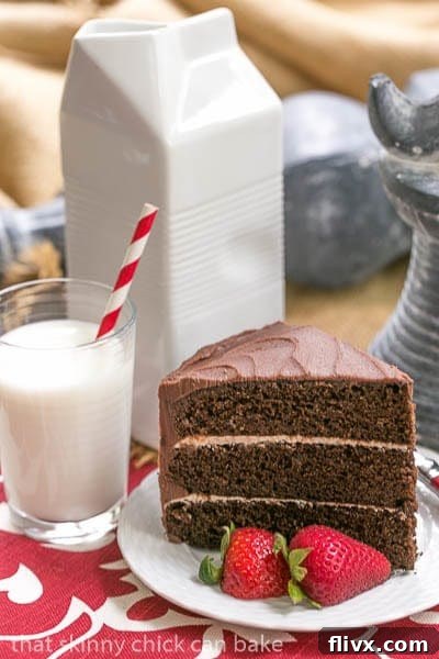 A perfectly portioned slice of triple-layer chocolate cake with generous vanilla buttercream on a white plate, accompanied by a tall glass of cold milk, ready to be enjoyed.