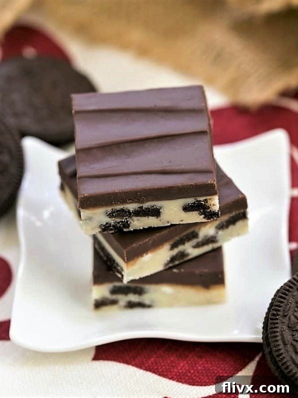 Several pieces of Double Decker Oreo Fudge are artfully stacked on a small white ceramic plate, surrounded by whole and broken Oreo cookies.
