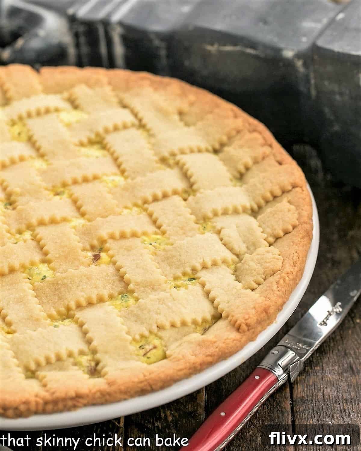 A close-up view of an uncut Pizza Rustica nestled in a white ceramic pie plate, with a rustic red-handled knife resting beside it, highlighting the intricate lattice top.