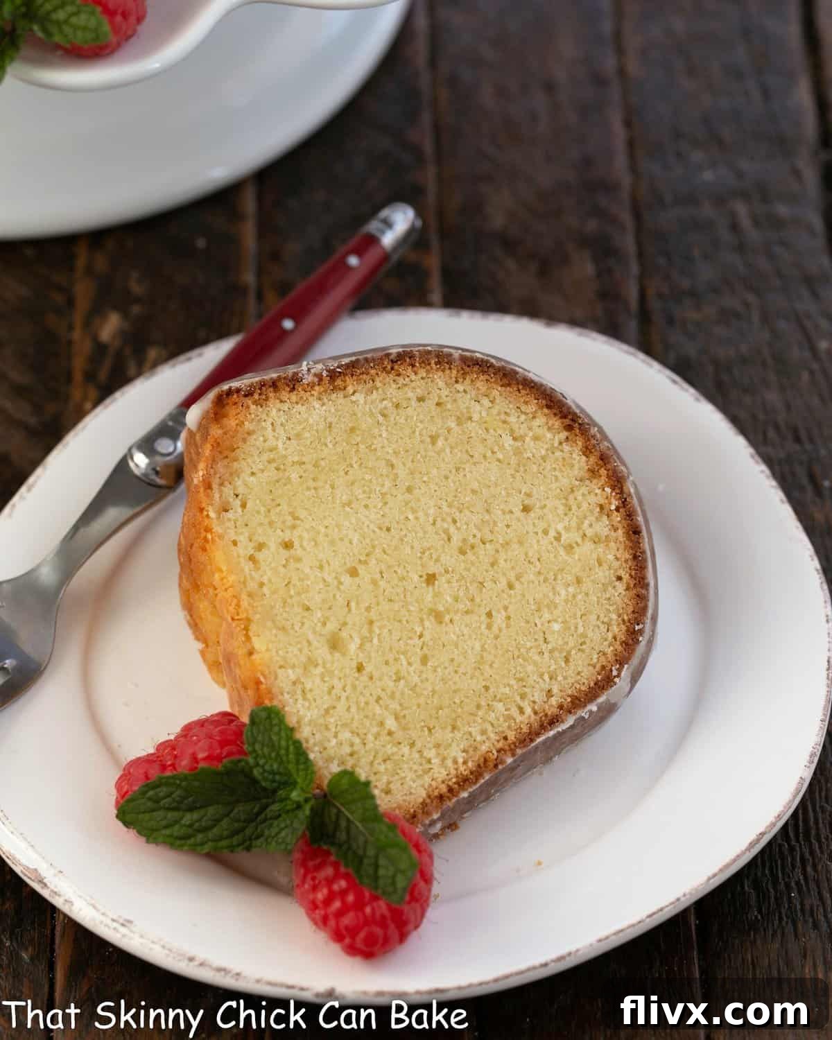 A delectable slice of sour cream pound cake on a pristine white dessert plate, accompanied by a elegant red-handled fork and a scattering of fresh, juicy raspberries.