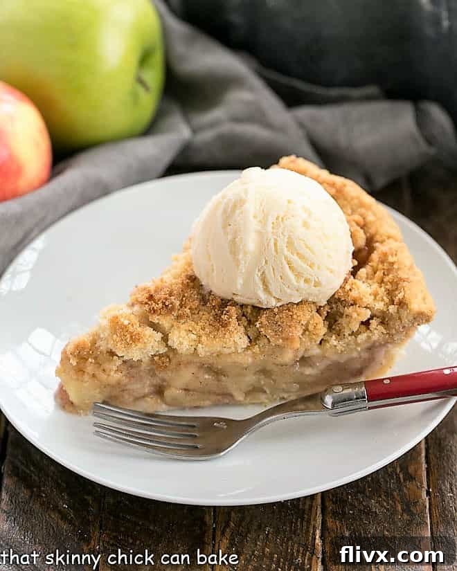 A perfectly baked slice of Dutch Apple Pie, generously topped with a scoop of creamy vanilla ice cream, presented on a pristine white dessert plate. The golden-brown streusel and tender apple filling are visibly enticing.