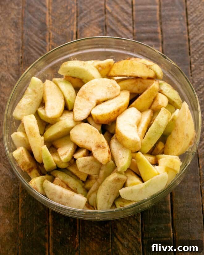 Close-up shot of apple slices being mixed with granulated sugar, fresh lemon juice, cinnamon, and nutmeg in a large bowl, preparing them for the pie filling.