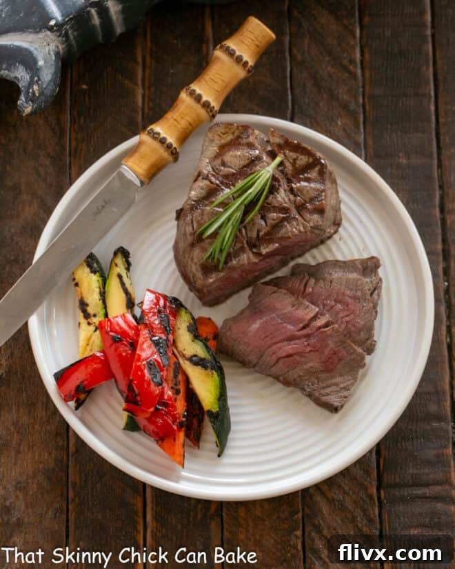 Perfectly sliced Filet Mignon with a glistening Red Wine Balsamic Sauce, served on a white plate alongside vibrant green vegetables and a steak knife.