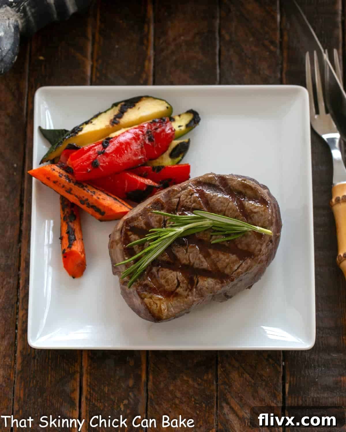 An overhead view of a beautifully grilled filet mignon, garnished with rosemary, on a square white plate, accompanied by perfectly grilled vegetables.
