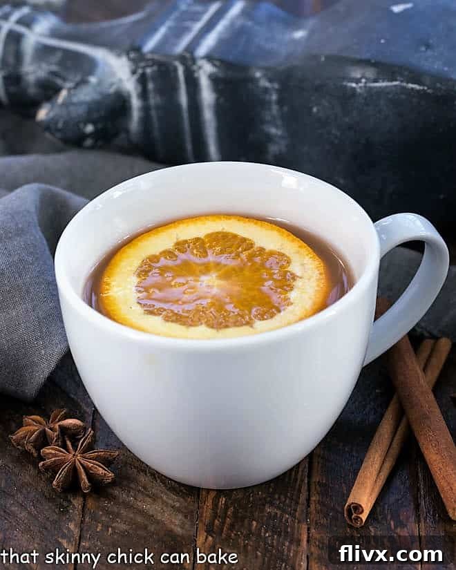 A charming round white mug filled with steaming Mulled Apple Cider, artfully topped with a fresh orange slice and a cinnamon stick for garnish.