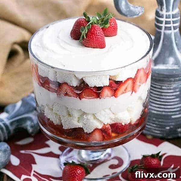 A glistening Strawberry Cheesecake Trifle in an elegant glass trifle bowl, adorned with three whole, ripe strawberries on top.