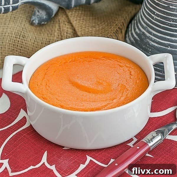 A round white baking dish filled with golden Carrot Souffle, garnished with fresh herbs, alongside a red handled serving spoon. Perfect for a holiday spread.