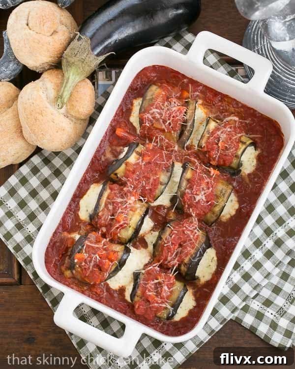 Eggplant Rollatini in a white casserole dish surrounded by wheat rolls and a whole eggplant