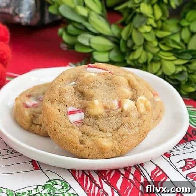 White Chocolate Candy Cane Cookies - A chewy, buttery cookie filled with white chocolate chips and crushed candy canes Three White Chocolate Candy Cane Cookies arranged on a white plate, nestled on a festive holiday napkin. The cookies are golden brown with visible white chocolate chips and crushed red and white candy cane pieces.