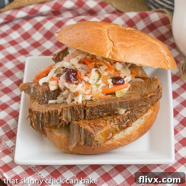 Oven Braised Texas Brisket sandwich topped with coleslaw sits on a white plate with a red and white gingham tablecloth underneath
