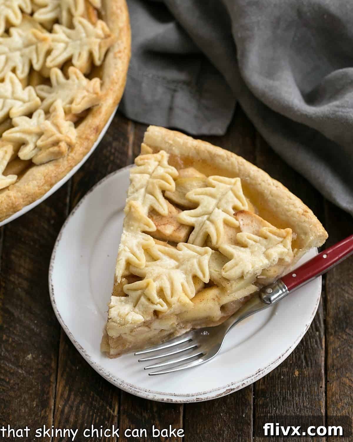 Overhead of a slice of apple pie on a round white dessert plate next to a partial view of the rest of the pie, showcasing the golden crust and juicy filling.