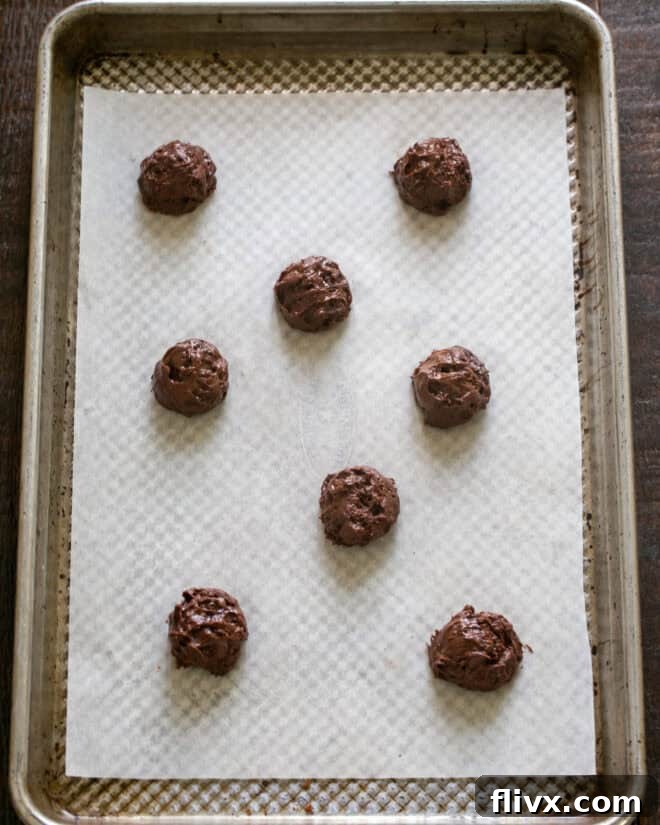 Teaspoonfuls of sticky brownie cookie batter being scooped onto a prepared baking sheet.