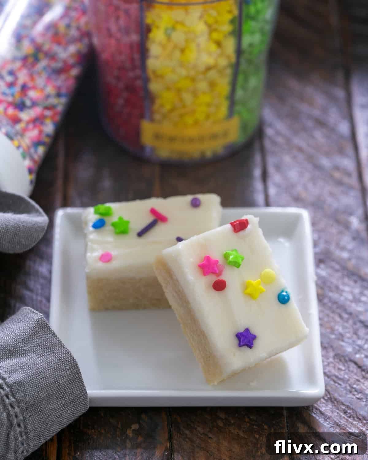 Two decadently frosted sugar cookie bars, adorned with colorful sprinkles, resting on a small white plate.