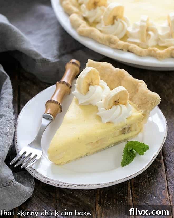 A perfectly cut slice of banana cream pie on a white plate with a bamboo handle fork, showcasing its layers of crust, custard, bananas, and whipped cream. The whole pie is visible in the background.