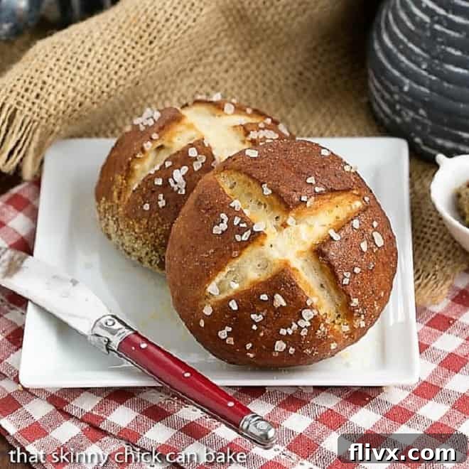 Two perfectly baked homemade pretzel rolls on a square white plate, accompanied by a red-handled knife. The rolls boast a deep, glossy brown crust and a classic 'X' score, inviting a delicious bite.