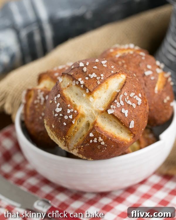 A beautiful white bowl brimming with freshly baked, perfectly golden-brown homemade pretzel rolls, ready for serving. Their glossy, salted crusts are prominent.