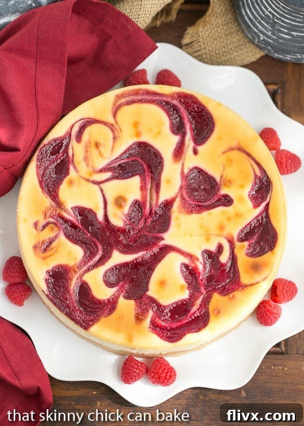An overhead view of the entire White Chocolate Raspberry Swirl Cheesecake on a elegant white serving plate, surrounded by fresh raspberries, showing the full beauty of the swirl.