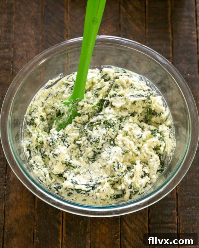 A hand mixing the spinach and cheese filling in a bowl, next to a platter of cooked jumbo pasta shells.