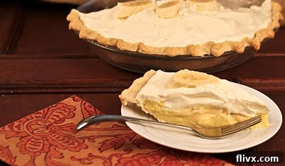 A perfect slice of banana cream pie on a dessert plate, with the full pie blurred in the background, inviting a delicious treat