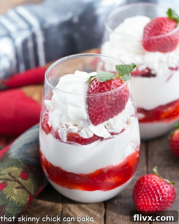 A close-up of two elegant parfait glasses showcasing layered fruit desserts with whipped cream, crushed meringue cookies, and vibrant sliced strawberries, with fresh strawberries scattered nearby.