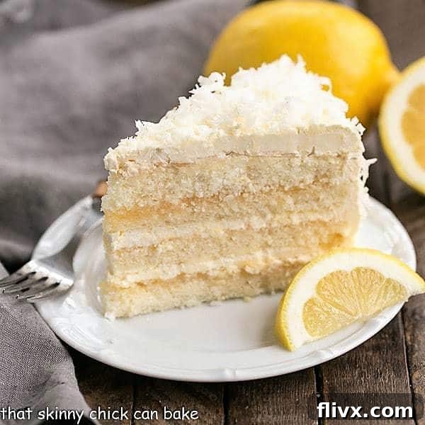 A close-up of a slice of Lemon Layer Cake, beautifully frosted and garnished with a lemon wedge, on a white ceramic plate.