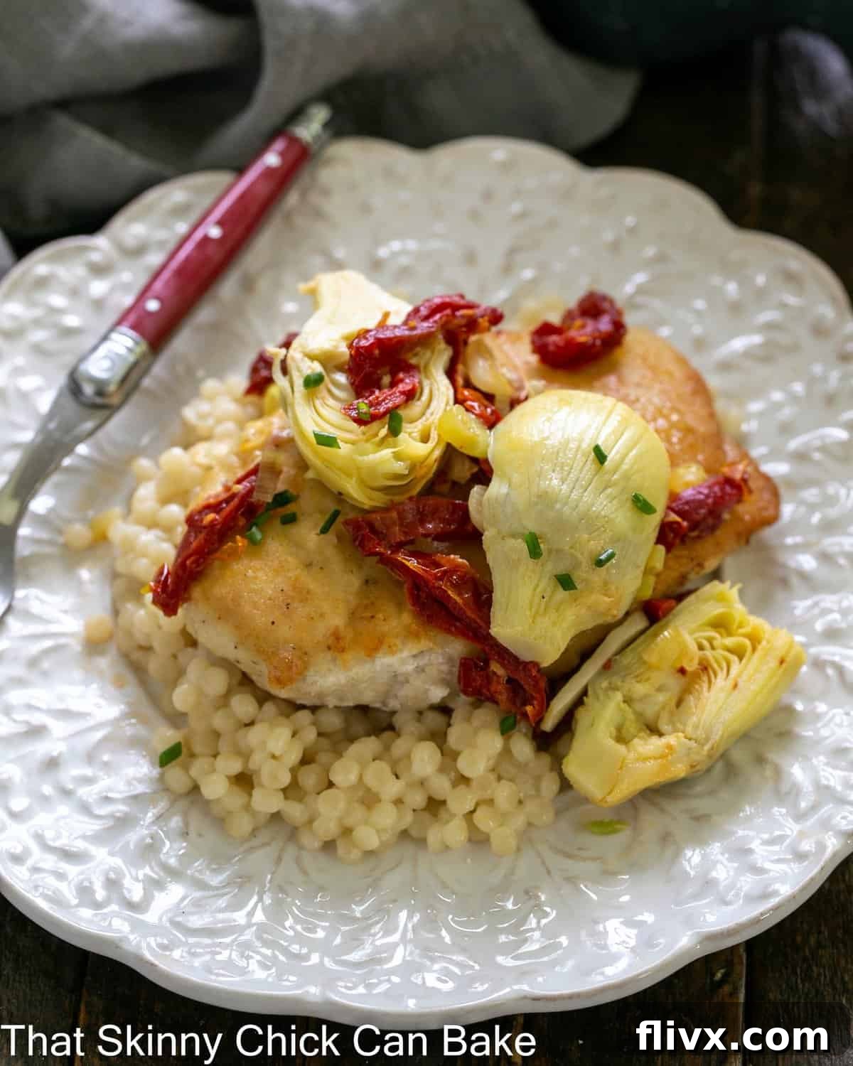 close overhead view of chicken with artichokes and sundried tomatoes over couscous on a white plate with a red handled fork.