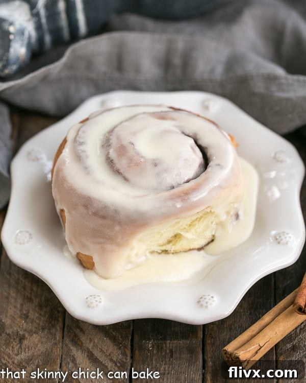 A close up of a perfectly glazed homemade cinnamon bun on a decorative white plate, showing the swirled cinnamon filling.