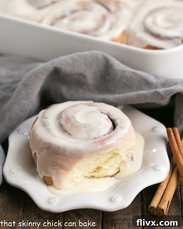 A glazed cinnamon bun on a white plate with a baking pan filled with more cinnamon rolls blurred in the background.