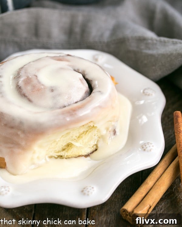 A beautifully glazed cinnamon bun garnished with cinnamon sticks on a white plate, ready to be enjoyed.
