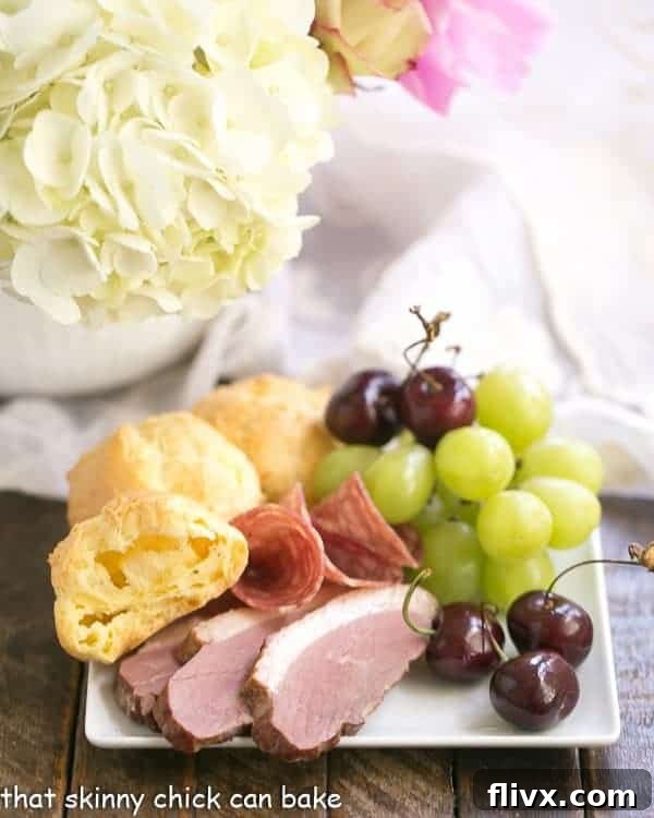 A close-up shot of Triple Cheese Gougeres artfully arranged on a rustic plate, surrounded by a vibrant charcuterie assortment including cured meats, fresh fruit, and olives, emphasizing their role in a gourmet spread.